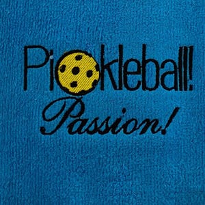 May include: A blue towel with the text "Pickleball! Passion!" embroidered in black. A yellow pickleball with black holes is embroidered next to the text.