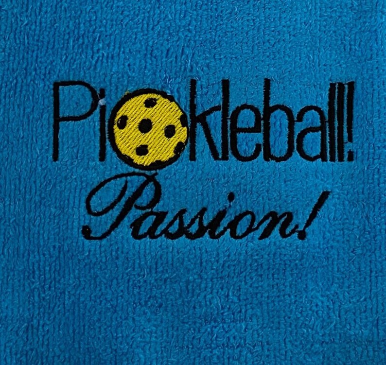 May include: A blue towel with the text "Pickleball! Passion!" embroidered in black. A yellow pickleball with black holes is embroidered next to the text.
