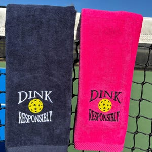 Op de afbeelding: Twee handdoeken, een donkerblauwe en een roze, met de tekst "DINK RESPONSIBLY" en een geborduurde gele pickleball-afbeelding op elk.
