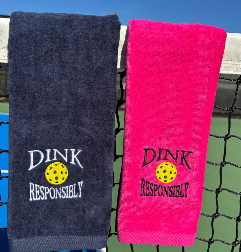 Op de afbeelding: Twee handdoeken, een donkerblauwe en een roze, met de tekst "DINK RESPONSIBLY" en een geborduurde gele pickleball-afbeelding op elk.