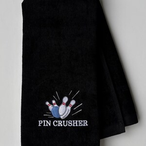 May include: A black hand towel featuring an embroidered graphic of a blue bowling ball striking white bowling pins with red stripes. The text PIN CRUSHER is embroidered in white capital letters below the bowling graphic on the fabric.