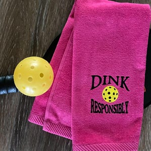 Op de afbeelding: Roze handdoek met de tekst "DINK RESPONSIBLY" erop geborduurd, met een gele pickleball in het midden van de tekst.