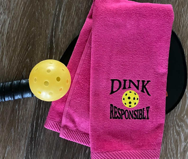 Op de afbeelding: Roze handdoek met de tekst "DINK RESPONSIBLY" erop geborduurd, met een gele pickleball in het midden van de tekst.