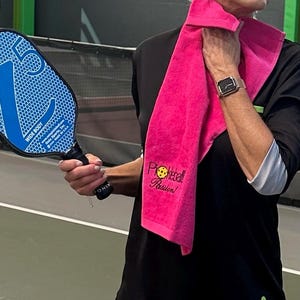 May include: A woman wearing a black shirt with a pink towel around her neck. The towel has the text "Pickleball Passion!" embroidered on it. She is holding a blue pickleball paddle.