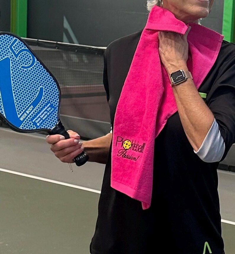 May include: A woman wearing a black shirt with a pink towel around her neck. The towel has the text "Pickleball Passion!" embroidered on it. She is holding a blue pickleball paddle.