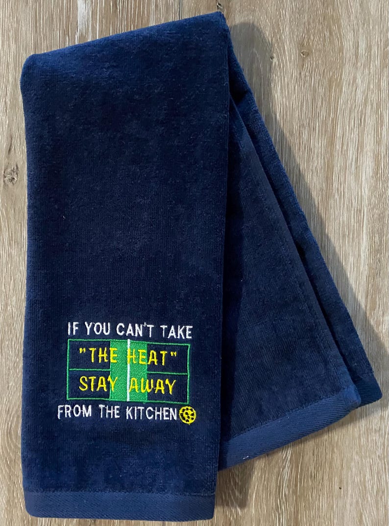 May include: A dark navy blue kitchen towel with embroidered text. The text reads: 'If you can't take 'the heat' stay away from the kitchen.'  The embroidered text is in yellow and green, resembling a scoreboard. The towel is folded in half, showing both sides. The towel is made of a soft, plush material.