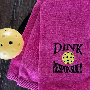 Op de afbeelding: Roze handdoek met zwarte borduurwerk die "Dink Responsibly" zegt met een gele en zwarte pickleball-afbeelding.