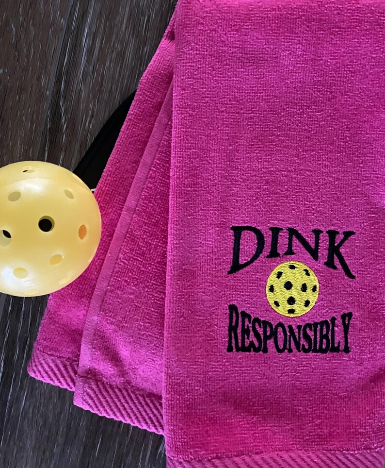 Op de afbeelding: Roze handdoek met zwarte borduurwerk die "Dink Responsibly" zegt met een gele en zwarte pickleball-afbeelding.