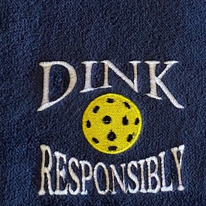 Op de afbeelding: Een donkerblauwe handdoek met witte borduurwerk die "DINK RESPONSIBLY" zegt en een gele en zwarte pickleball.