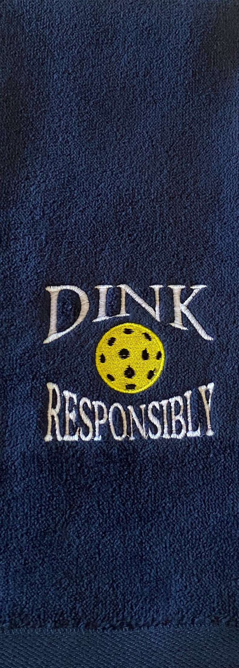 Op de afbeelding: Een donkerblauwe handdoek met witte borduurwerk die "DINK RESPONSIBLY" zegt en een gele en zwarte pickleball.