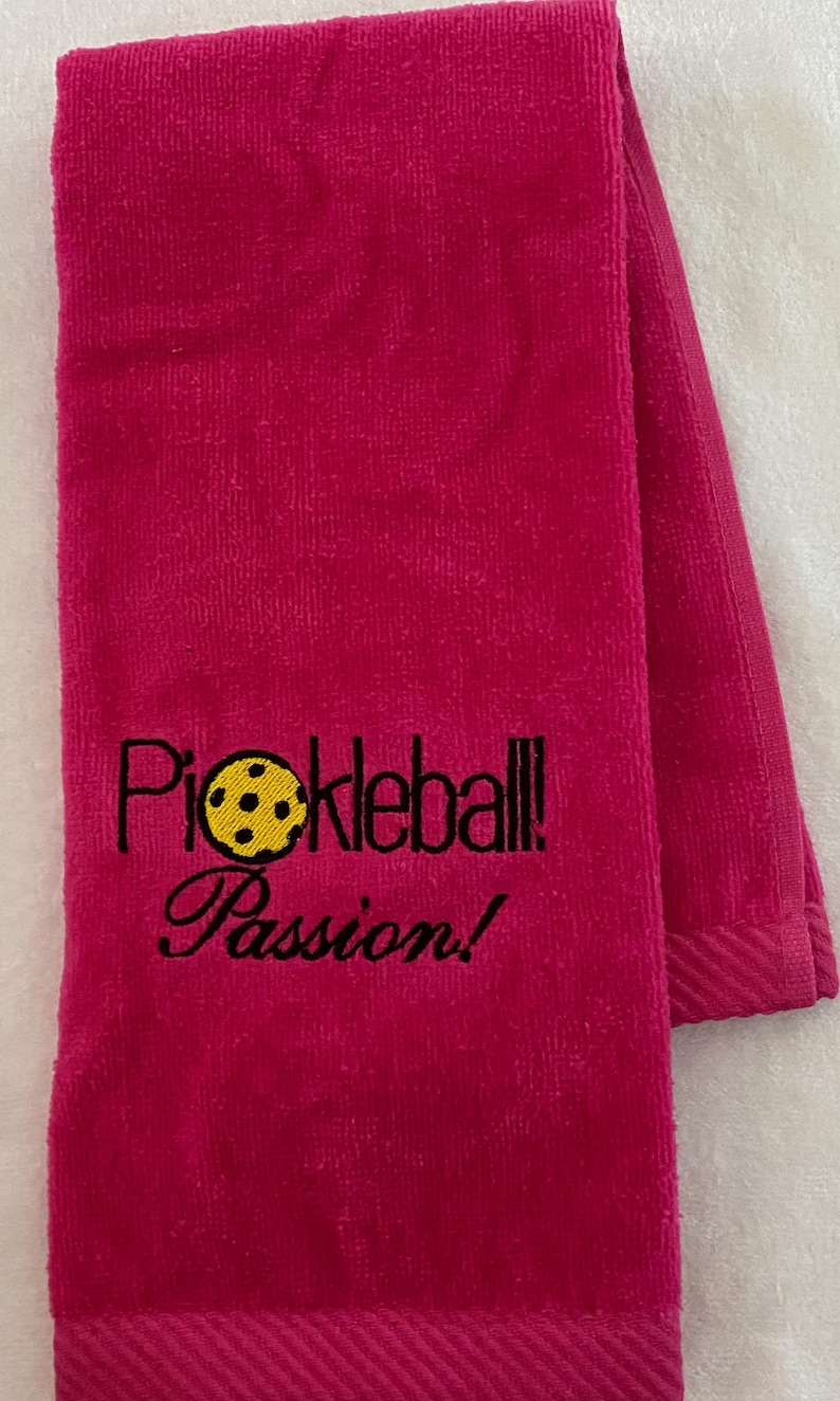 May include: A pink towel with the text "Pickleball Passion!" embroidered in black with a yellow pickleball graphic.