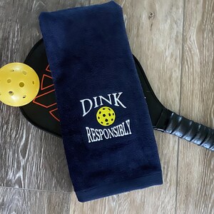 Op de afbeelding: Een donkerblauwe handdoek met de tekst "DINK RESPONSIBLY" geborduurd in wit. Op de achtergrond zijn een gele pickleball en een zwarte pickleball-paddle te zien.