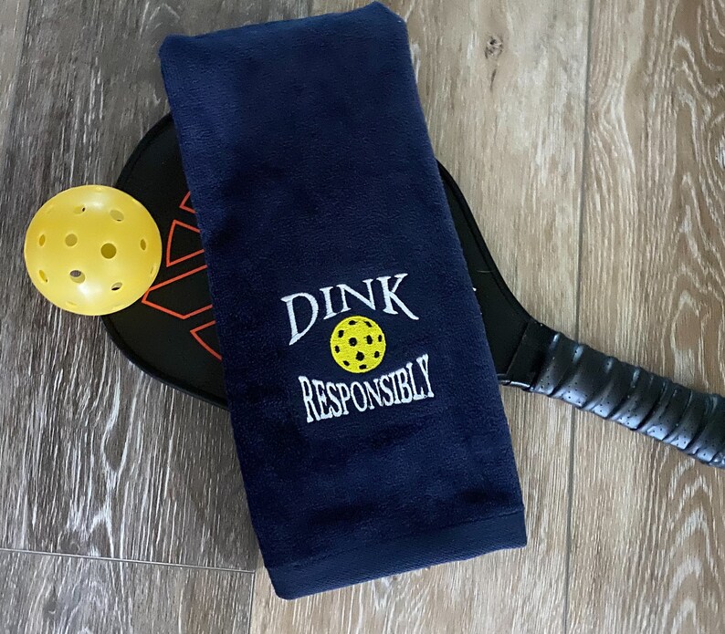 Op de afbeelding: Een donkerblauwe handdoek met de tekst "DINK RESPONSIBLY" geborduurd in wit. Op de achtergrond zijn een gele pickleball en een zwarte pickleball-paddle te zien.