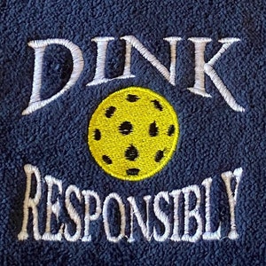 Op de afbeelding: Een marineblauwe fleece handdoek met witte borduurwerk dat "DINK RESPONSIBLY" zegt en een geborduurde gele pickleball.