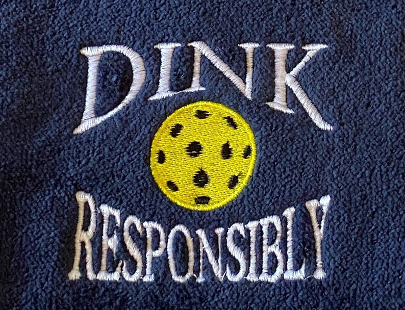 Op de afbeelding: Een marineblauwe fleece handdoek met witte borduurwerk dat "DINK RESPONSIBLY" zegt en een geborduurde gele pickleball.