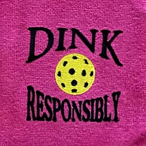 Op de afbeelding: Roze handdoek met zwarte borduurwerk die "DINK RESPONSIBLY" zegt met een gele en zwarte pickleball in het midden.