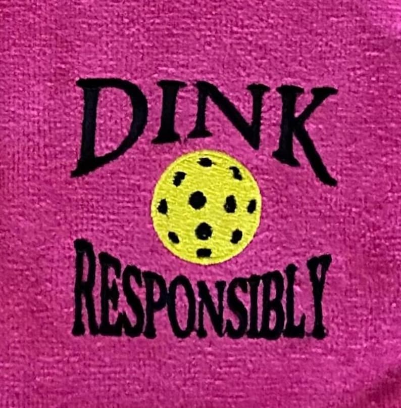 Op de afbeelding: Roze handdoek met zwarte borduurwerk die "DINK RESPONSIBLY" zegt met een gele en zwarte pickleball in het midden.