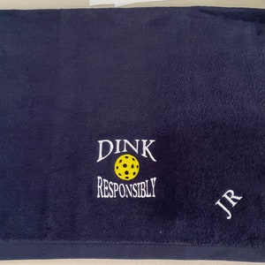 Op de afbeelding: Een donkerblauwe handdoek met witte borduurwerk die "DINK RESPONSIBLY" en een gele pickleball zegt. De handdoek heeft ook de initialen "JR" geborduurd in wit.
