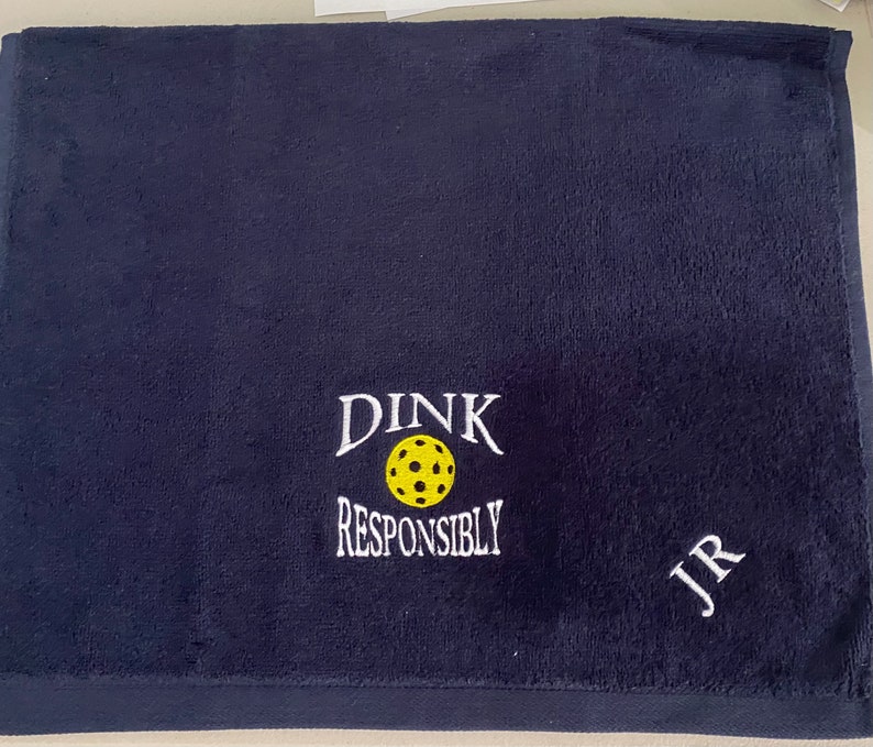 Op de afbeelding: Een donkerblauwe handdoek met witte borduurwerk die "DINK RESPONSIBLY" en een gele pickleball zegt. De handdoek heeft ook de initialen "JR" geborduurd in wit.
