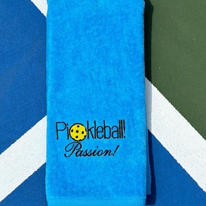 May include: A bright blue towel with the words "Pickleball! Passion!" embroidered in black with a yellow pickleball graphic.