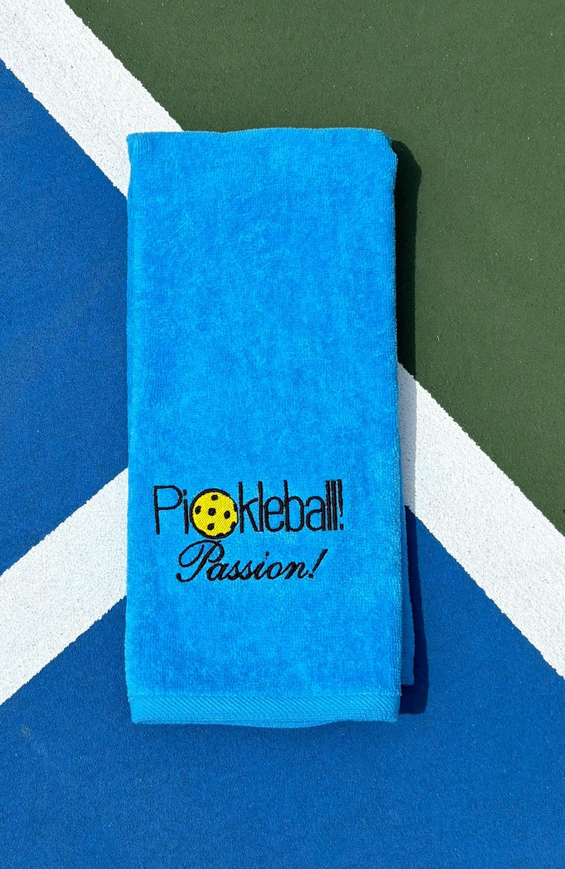 May include: A bright blue towel with the words "Pickleball! Passion!" embroidered in black with a yellow pickleball graphic.