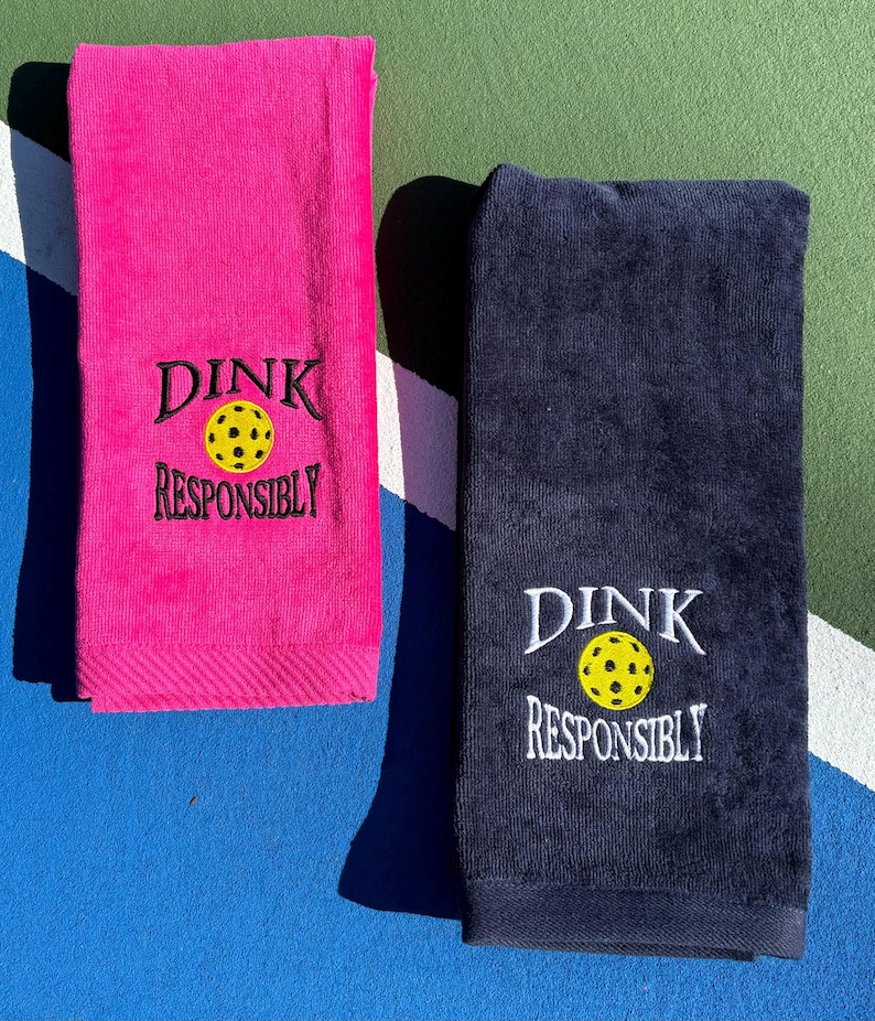 Op de afbeelding: Twee handdoeken met de tekst "Dink Responsibly" en een gele pickleball-afbeelding. Een handdoek is roze en de andere is donkerblauw.