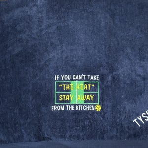 K&ouml;nnte beinhalten: Ein dunkelblaues Handtuch mit dem Text "If you can't take "the heat" stay away from the kitchen" in gr&uuml;n und gelb gestickt. Das Handtuch hat auch das Wort "TYSON" in wei&szlig; gestickt.