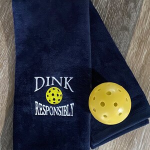Op de afbeelding: Een donkerblauwe handdoek met witte borduurwerk met de tekst "Dink Responsibly" en een gele pickleball.