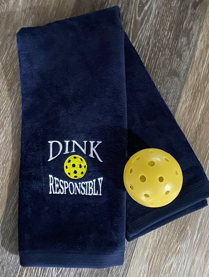 Op de afbeelding: Een donkerblauwe handdoek met witte borduurwerk met de tekst "Dink Responsibly" en een gele pickleball.