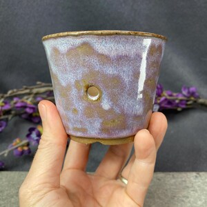 Peut inclure: Un petit pot en céramique conique présentant un émail tacheté dans des tons de lavande, de blanc et de brun terreux. Le récipient a une finition lisse et brillante et comprend un seul trou de drainage circulaire sur le côté du corps.
