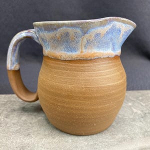 Peut inclure: Un pichet en céramique brun avec un glaçage bleu et blanc sur le bord supérieur. Le pichet a un corps arrondi et une anse.