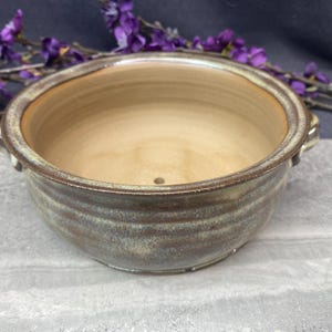 Puede incluir: Un cuenco de cerámica redondo con dos pequeñas asas laterales, que presenta un interior liso de color beige claro y un exterior texturizado con un esmalte moteado en tonos marrón, gris y verde apagado, con un pequeño orificio de drenaje.