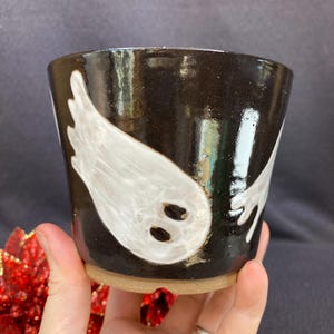 May include: A ceramic cup with a glossy black glaze featuring a hand-painted white ghost design. The ghost has two dark oval eyes and a trailing body shape. The cup has a wide, open top and a natural, unglazed tan clay base at the bottom.