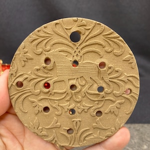 May include: A circular tan-colored disc featuring an embossed horse silhouette surrounded by intricate scrolling leaf patterns. The disc contains several small circular cutouts throughout the design, revealing glimpses of red accents behind the surface.