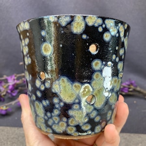 May include: A cylindrical ceramic planter featuring a glossy dark navy blue glaze with scattered light green and cream-colored crystalline spots. The vessel has several circular drainage holes distributed across its sides and a plain clay base.