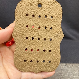 May include: A rectangular tan clay display board with a scalloped edge and an intricate embossed scrollwork pattern. The board features a single hole at the top for hanging and fifteen smaller circular holes arranged in five rows of three.