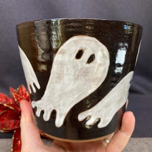 May include: A cylindrical ceramic planter with a glossy black glaze featuring hand-painted white ghost figures with oval eyes and scalloped bottom edges. The pot has a smooth, reflective surface and a wide, open top rim for holding small plants.
