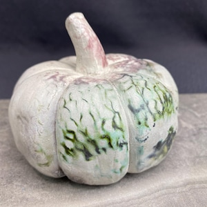 May include: A white ceramic pumpkin with green and pink glaze. The pumpkin has a textured surface and a slightly uneven shape.
