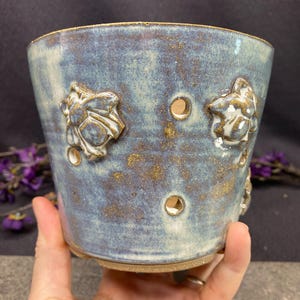 May include: A ceramic planter with a mottled blue and brown glaze, featuring raised floral relief decorations and several circular drainage holes distributed across the exterior surface. The pot has a wide, open top and a sturdy base.