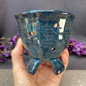 May include: A small, round, blue ceramic planter with a glossy, mottled finish. The pot features three short, sturdy legs and several circular drainage holes distributed around the upper body, showcasing a deep indigo and teal color palette.