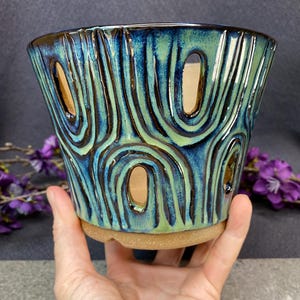 May include: A ceramic planter featuring a glossy glaze in shades of deep blue and light green. The exterior has a textured, swirling pattern with several vertical cutouts throughout the body. The base is made of unglazed, speckled tan clay.