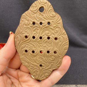 May include: A tan, shield-shaped ceramic ornament featuring an intricate, embossed swirling vine pattern across its surface. The piece includes a single hole at the top for hanging and twelve smaller, evenly spaced circular perforations.