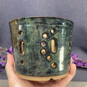 May include: A cylindrical ceramic planter with a glossy, mottled dark green and black glaze. The exterior features numerous circular and elongated vertical cutouts arranged in a scattered pattern, revealing the light tan clay body underneath.