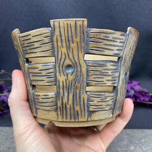May include: A handcrafted, ceramic planter with a rustic, wood-like design. The planter features horizontal slats and vertical supports, with a natural brown and dark brown color scheme. The open design allows for air circulation and drainage, perfect for small plants.