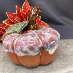 May include: A ceramic pumpkin figurine featuring a glossy brown base with a white and light blue drip glaze effect on the top. The pumpkin has a dark brown stem and a single green leaf attached to the top, with red poinsettia accents.