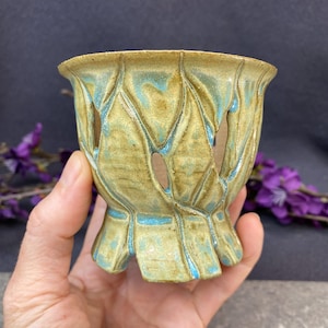 May include: A small ceramic vessel featuring a textured, leaf-like exterior design with a glossy finish. The surface displays a blend of muted olive green and vibrant turquoise blue glaze, with open cutouts between the raised leaf patterns.