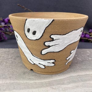 Puede incluir: Una maceta de cerámica de color marrón con un diseño de fantasmas blancos. Los fantasmas están pintados con ojos y contornos negros. La maceta tiene una forma ligeramente acampanada y es adecuada para uso en interiores o exteriores.
