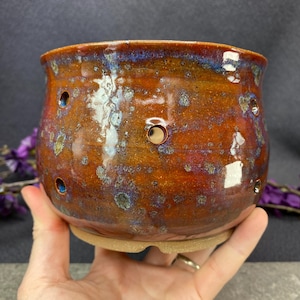 May include: A handcrafted ceramic pot with a glossy glaze in hues of brown, orange, and blue. The pot has several small holes around its body. It rests on a matching, unglazed base. Ideal for plants or decorative purposes.