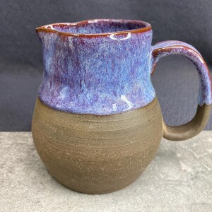 May include: A brown ceramic pitcher with a purple and brown glaze. The pitcher has a rounded body and a handle.