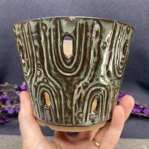 May include: A ceramic planter with a tapered cylindrical shape, featuring a dark brown and sage green glaze. The exterior is decorated with a repeating embossed arch pattern and includes several vertical cutouts for drainage and aeration.